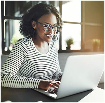 Smiling young African woman wearing glasses browsing online with a laptop while sitting alone at a table