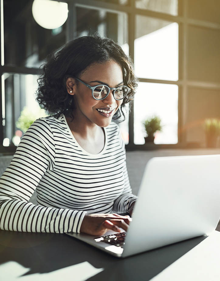Smiling young African woman wearing glasses browsing online with a laptop while sitting alone at a table