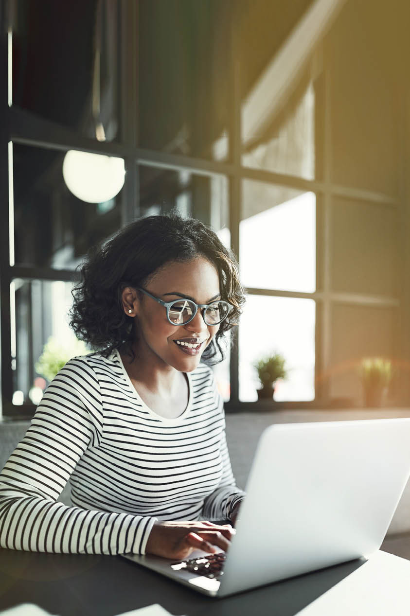 Smiling young African woman wearing glasses browsing online with a laptop while sitting alone at a table