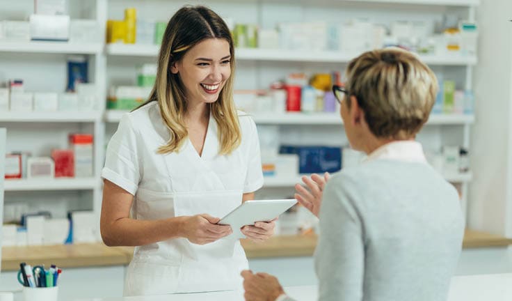 Female pharmacist selling medications at drugstore to a senior woman customer while using a tablet