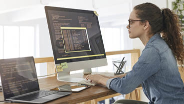 Side view portrait of female IT developer typing on keyboard with black and orange programming code on computer screen and laptop in contemporary office interior, copy space