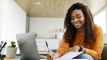 Business And Education Concept. Smiling young black woman sitting at desk working on laptop writing letter in paper notebook, free copy space. Happy millennial female studying using pc