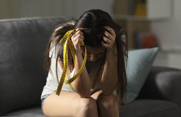 Sad teen worried about appearance holding a measure tape sitting on a couch in the living room at home