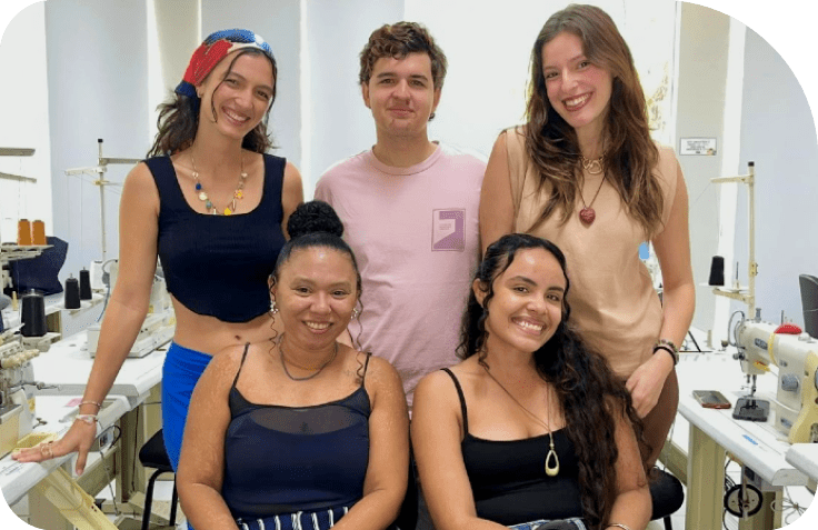 Cinco pessoas em roupas casuais est o em uma sala com v rias m quinas de costura, aparentando um ambiente de trabalho ou oficina de moda. Description generated by AI