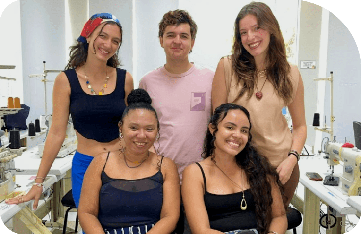 Cinco pessoas com roupas casuais est o em uma sala clara com v rias m quinas de costura ao redor, aparentando um ambiente de trabalho ou estudo relacionado  moda.  Description generated by AI