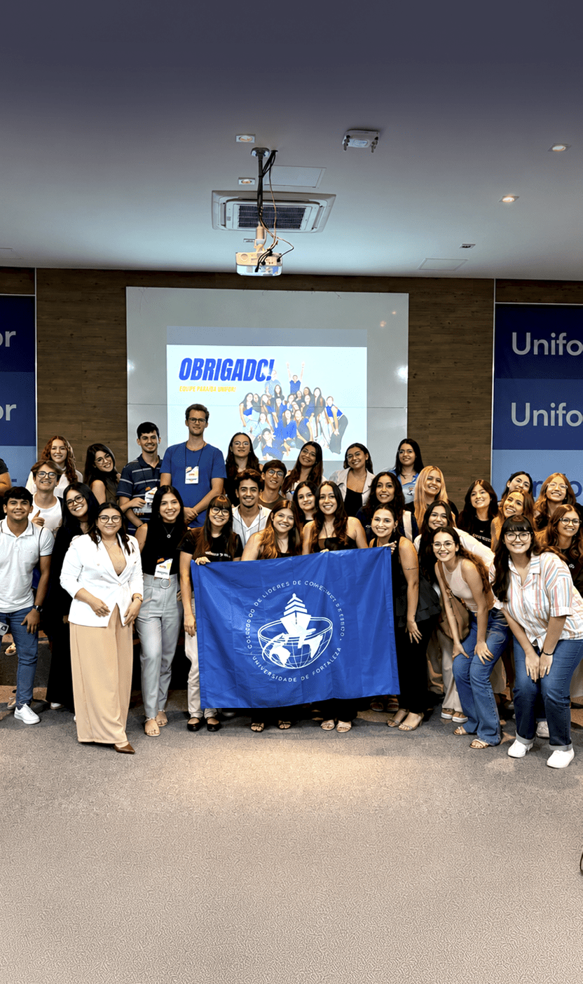 Um grupo grande de pessoas sorridentes est reunido em um ambiente interno segurando uma bandeira azul com um s mbolo branco enquanto atr s deles h  uma tela com a palavra “OBRIGADO!" e imagens de pessoas.  Description generated by AI