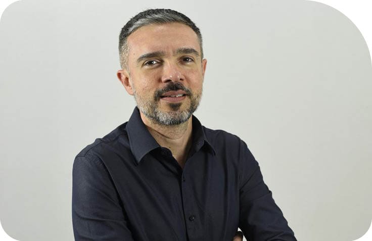 Pessoa adulta com cabelo curto e camisa escura de bot es est em p  com os bra os cruzados diante de um fundo claro.  Description generated by AI