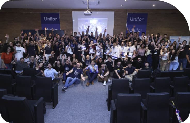 Grupo grande de pessoas reunidas em audit rio com cadeiras pretas, algumas sentadas e outras em p , sorrindo e acenando, com banners da Unifor ao fundo. Description generated by AI