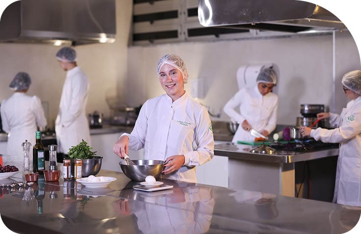 Pessoas vestindo jalecos e toucas trabalham em uma cozinha profissional preparando alimentos com utens lios e ingredientes sobre bancadas de inox. Description generated by AI