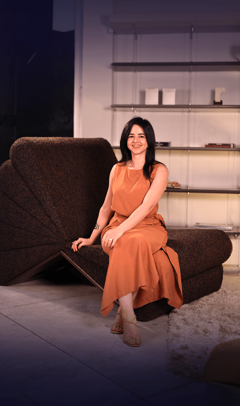 Pessoa com cabelo longo e liso usando vestido laranja sentada sorrindo em um sof escuro em ambiente interno com prateleiras ao fundo.  Description generated by AI