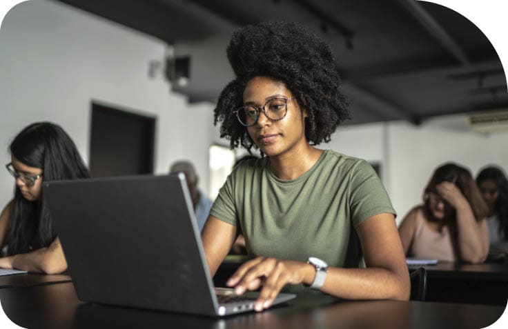 Pessoa com cabelo afro e culos usando camiseta verde est  concentrada trabalhando em um laptop em uma sala com outras pessoas ao fundo.  Description generated by AI