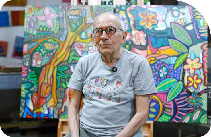 Pessoa idosa com culos e camiseta cinza sentada em frente a uma pintura colorida e vibrante com motivos florais e formas abstratas.  Description generated by AI