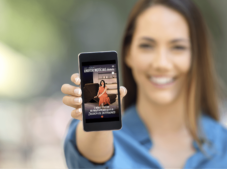 Pessoa sorridente segurando um celular com a tela mostrando a capa de uma revista digital chamada Unifor Not cias com uma pessoa sentada em um sof . Description generated by AI