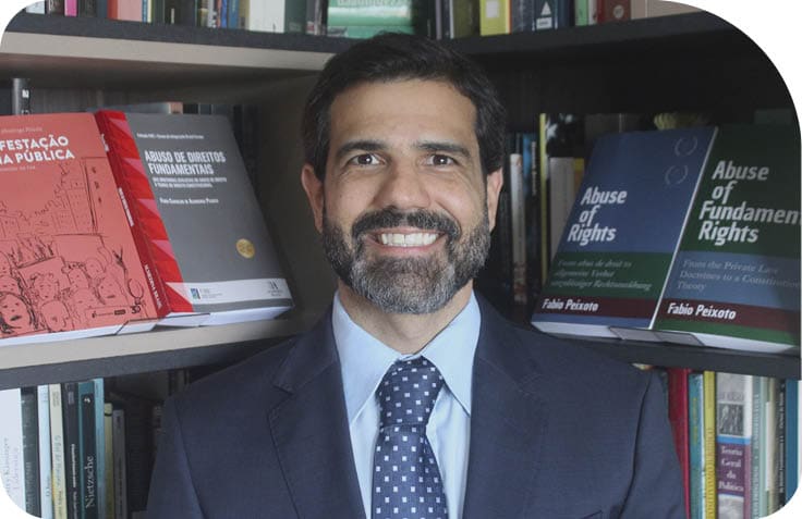 Pessoa adulta com barba sorrindo, vestindo terno e gravata, em frente a estante com livros sobre direitos fundamentais. Description generated by AI