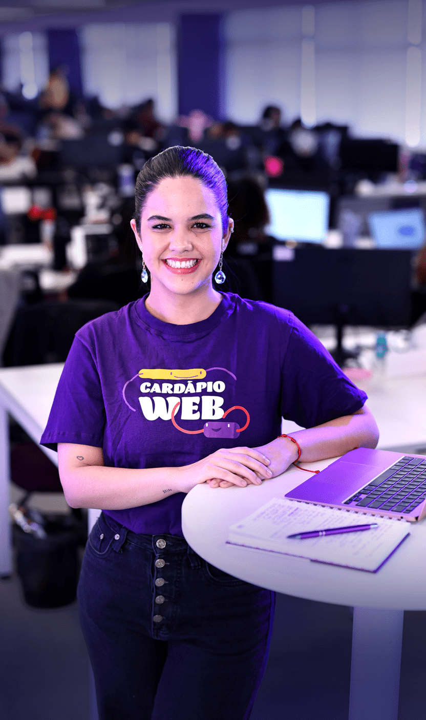 Pessoa sorridente com cabelo preso usando camiseta roxa com a frase “Card pio Web" em um ambiente de escrit rio moderno com computadores ao fundo. Description generated by AI