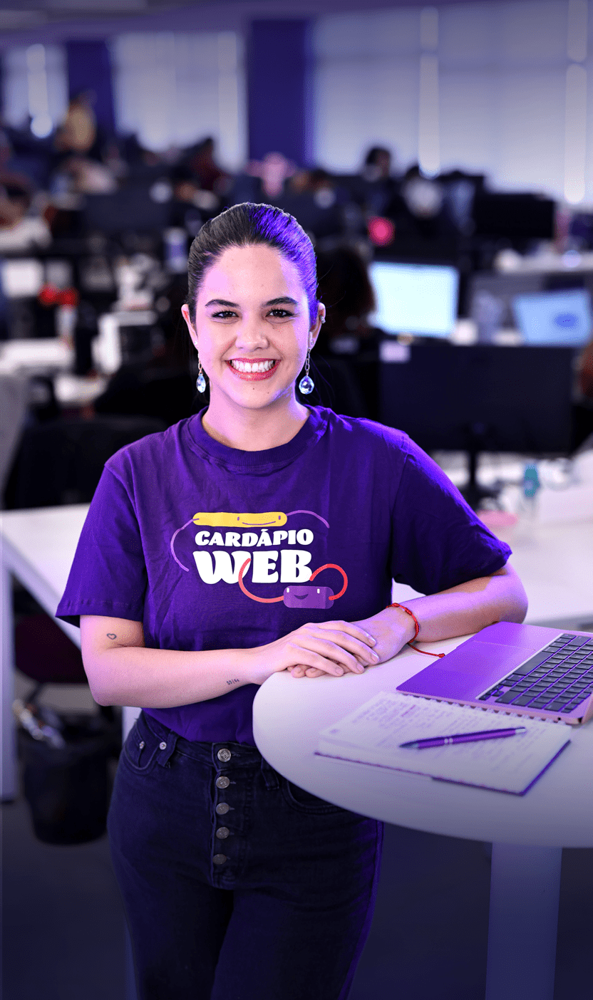 Pessoa adulta sorridente com cabelo preso usando camiseta roxa com a frase “Card pio Web" em ambiente de escrit rio com computadores ao fundo. Description generated by AI