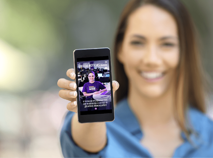 Pessoa sorridente segurando um celular com a capa de uma revista digital exibida na tela em um ambiente externo desfocado. Description generated by AI