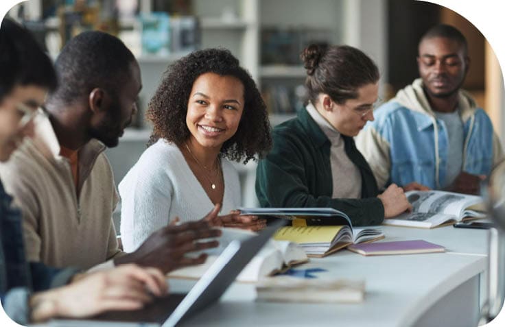 Quatro pessoas sentadas em uma mesa com livros e cadernos abertas, conversando e sorrindo em um ambiente de estudo ou trabalho. Description generated by AI