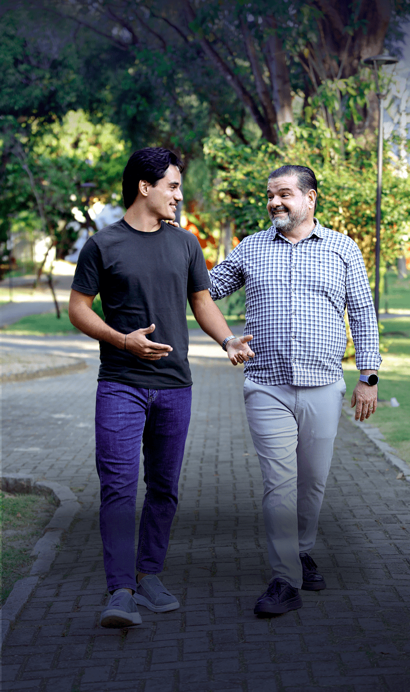 Duas pessoas caminhando juntas em um parque, sorrindo e conversando, uma vestindo camiseta preta e cal a azul e a outra camisa xadrez e cal a clara. Description generated by AI