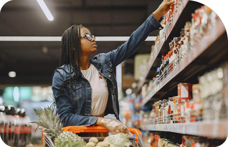 Pessoa com cabelo tran ado, culos e jaqueta jeans est  pegando um produto em uma prateleira de supermercado enquanto empurra um carrinho com frutas.  Description generated by AI