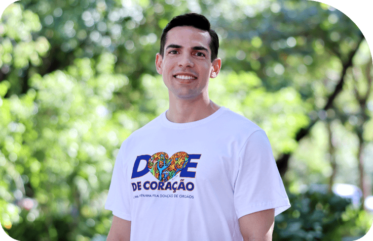 Pessoa sorridente vestindo camiseta branca com estampa colorida e texto sobre doa o de  rg os em ambiente externo com  rvores ao fundo.  Description generated by AI