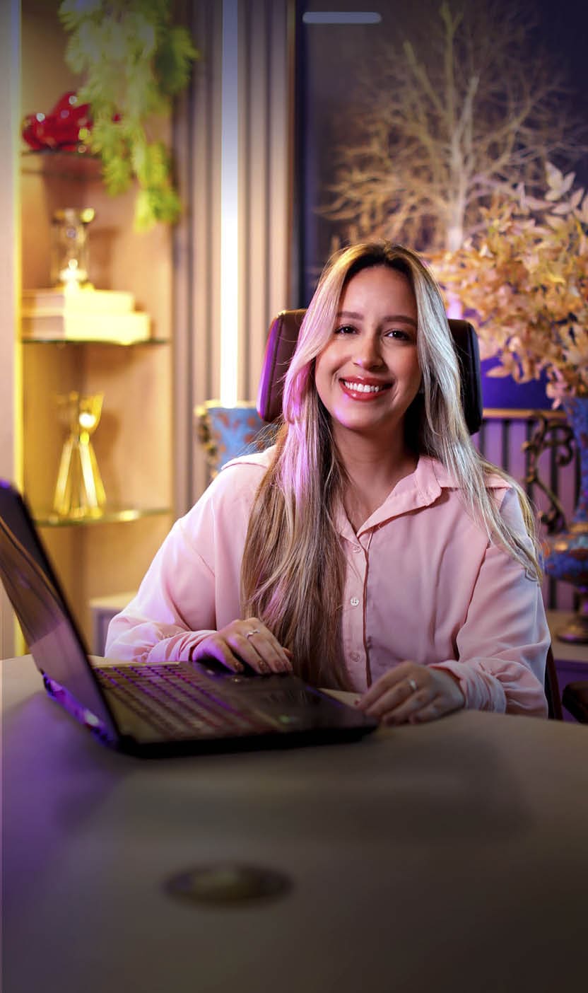 Pessoa adulta com cabelo longo e liso usando camisa rosa clara sorri enquanto est sentada em frente a um laptop em um ambiente interno decorado com plantas e ilumina  o quente.  Description generated by AI
