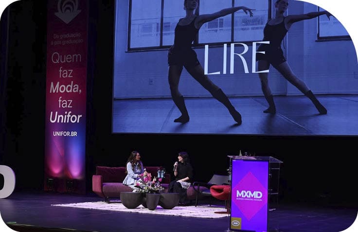 Duas pessoas sentadas em um palco decorado com flores e m veis modernos participam de uma conversa durante um evento com pain is e ilumina o colorida.  Description generated by AI