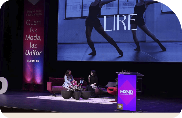 Duas pessoas sentadas em um palco conversando em um ambiente de evento com ilumina o roxa e telas ao fundo exibindo as palavras “LIRE" e "MXMD".  Description generated by AI