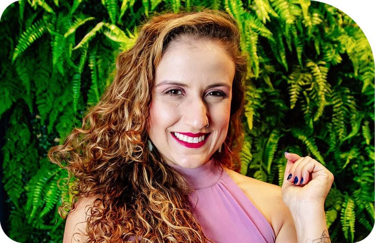 Pessoa com cabelo cacheado e longo usando batom vermelho e vestido rosa claro sorrindo em frente a um fundo de folhagens verdes. Description generated by AI