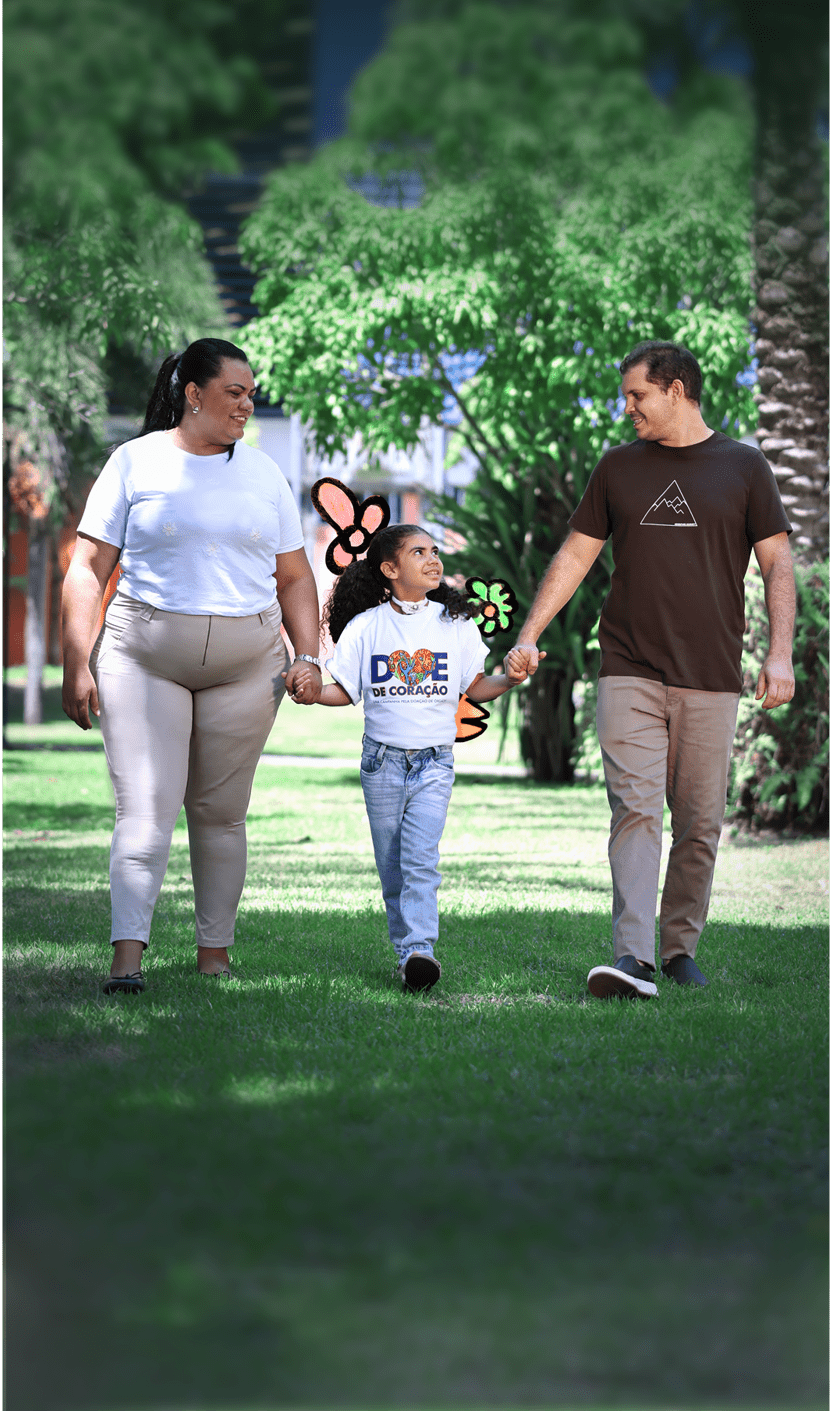 Tr s pessoas caminhando de m os dadas em um parque ensolarado com rvores ao fundo, sendo uma crian a no centro usando camiseta branca com estampa colorida e duas pessoas adultas ao lado vestindo roupas casuais.  Description generated by AI