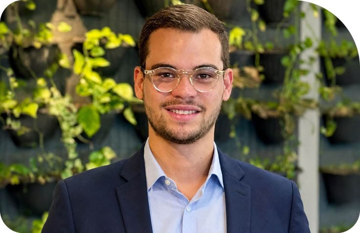 Pessoa adulta usando culos, camisa azul clara e blazer azul escuro sorrindo em ambiente com plantas ao fundo.  Description generated by AI