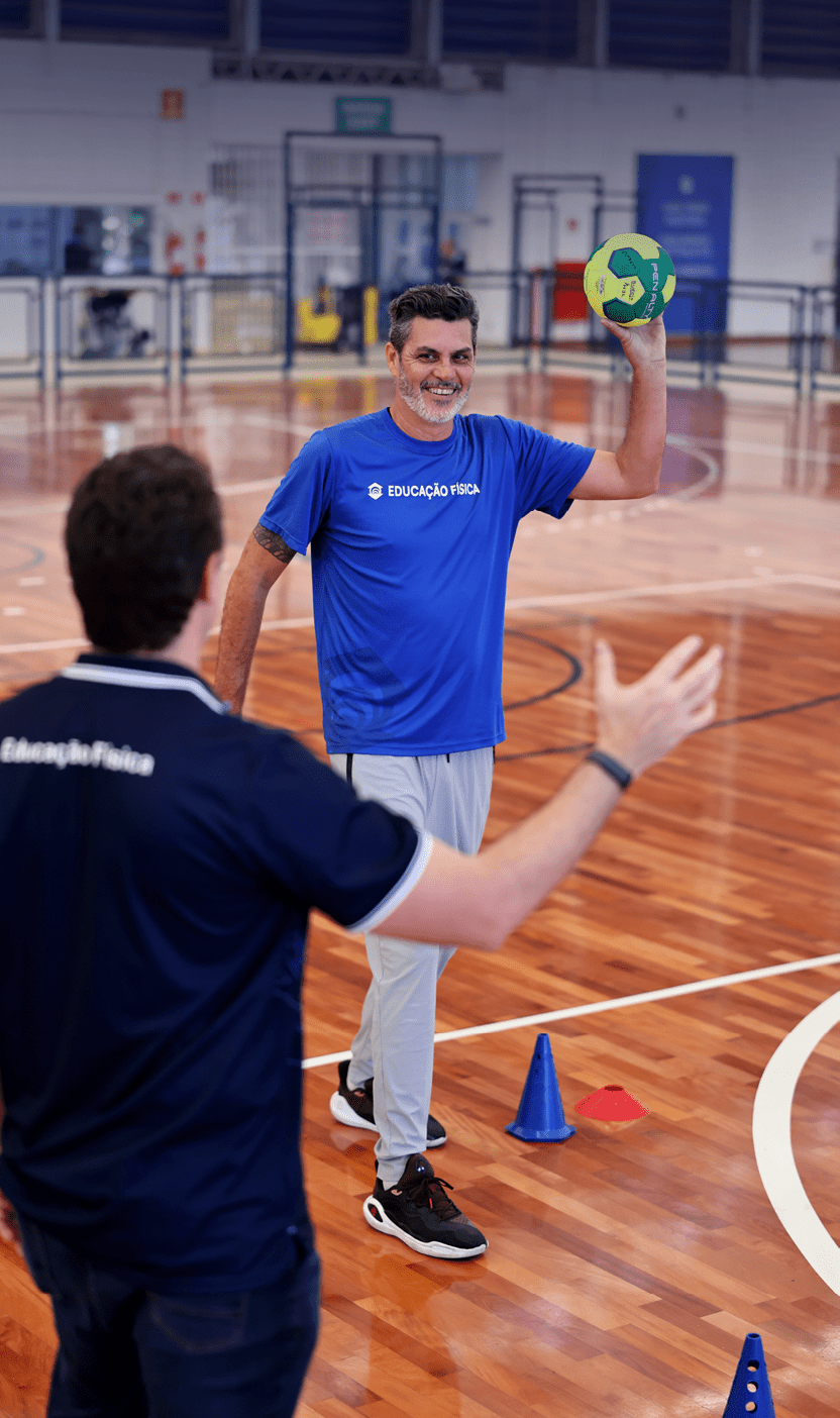 Duas pessoas em uma quadra esportiva interna, uma segurando uma bola de handebol e sorrindo enquanto a outra est de costas com os bra os abertos.  Description generated by AI