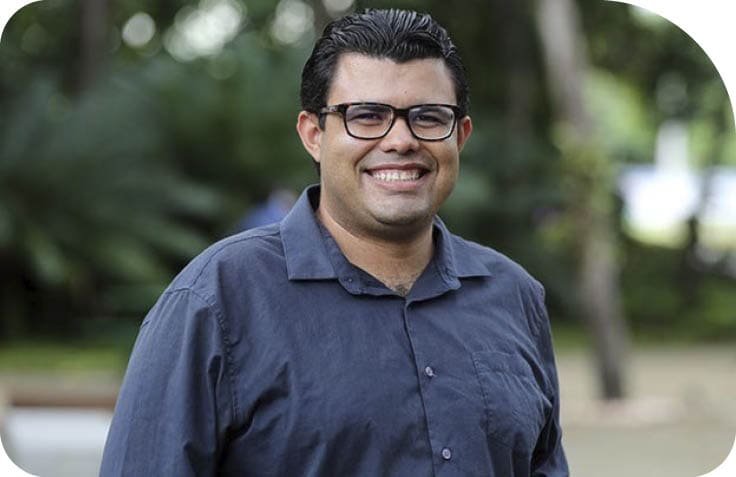 Pessoa adulta sorridente usando culos e camisa azul escura em ambiente externo com  rvores ao fundo.  Description generated by AI