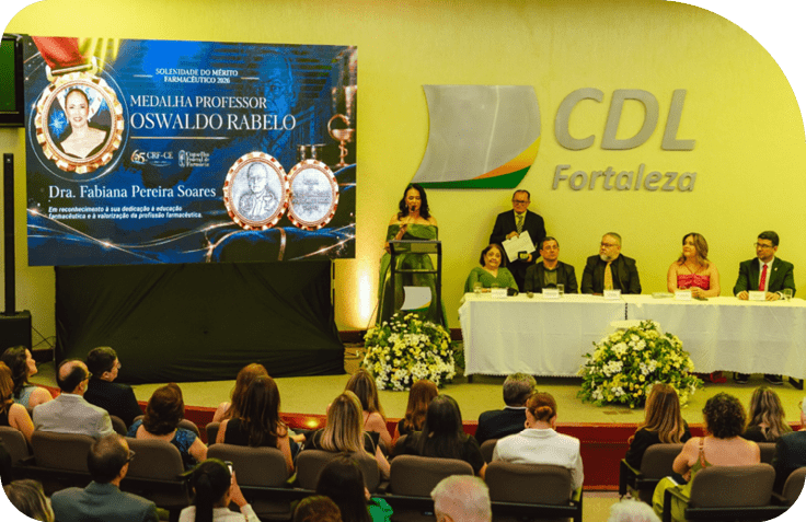 Evento formal em audit rio com pessoas sentadas em mesa principal decorada com arranjos florais e uma pessoa falando ao microfone ao lado de um tel o exibindo homenagem a professor Oswaldo Rabelo. Description generated by AI