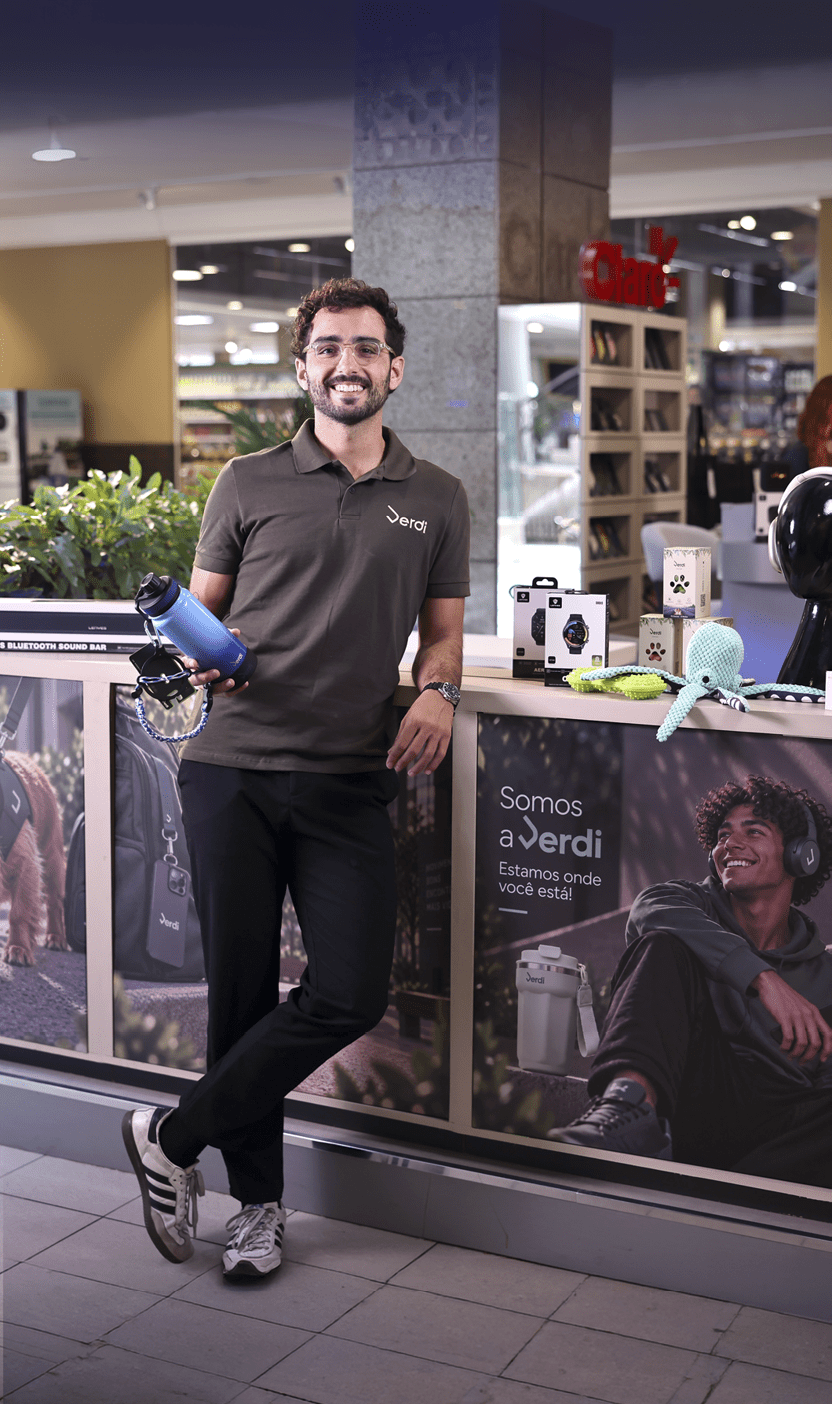 Pessoa adulta com cabelo encaracolado e culos sorri enquanto segura um microfone azul em uma loja com produtos eletr nicos e cartaz promocional ao fundo.  Description generated by AI