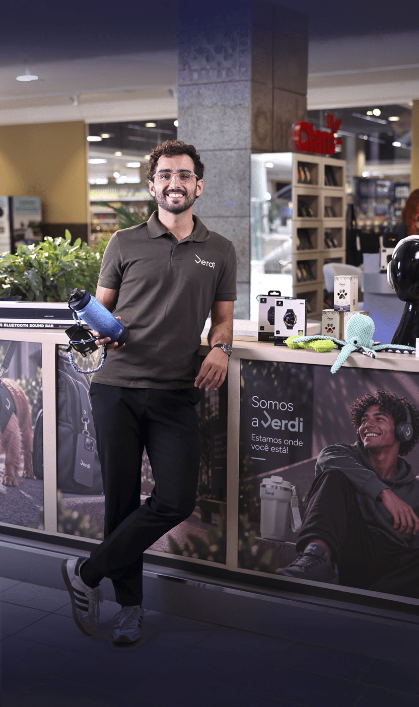 Pessoa adulta com camiseta polo escura e cal a preta sorri enquanto segura um secador de cabelo azul em um ambiente interno de loja com produtos eletr nicos expostos ao fundo. Description generated by AI