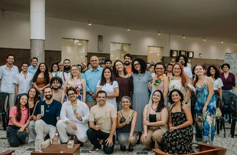Grupo grande de pessoas sorrindo e posando para foto em ambiente interno com cadeiras ao fundo, algumas segurando objetos coloridos. Description generated by AI