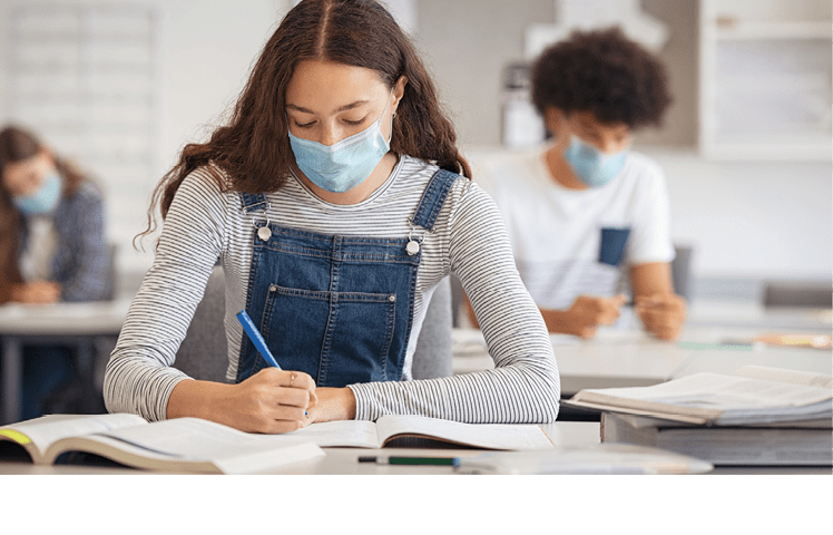 High school student taking notes from book while wearing face mask due to coronavirus emergency  Young woman sitting in class with their classmates and wearing surgical mask due to Covid-19 pandemic  Focused girl studying in classroom completing assignment during corona virus 